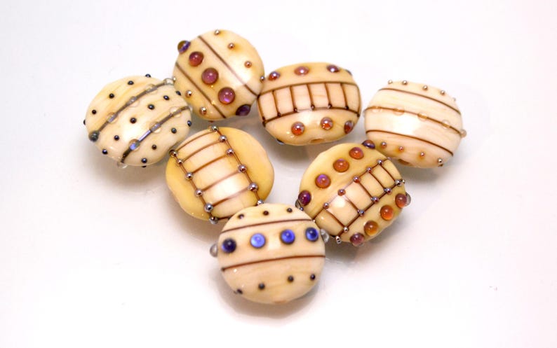 May include: A collection of eight glass beads with intricate patterns and colorful accents. The beads are cream-colored with brown, orange, and blue accents.