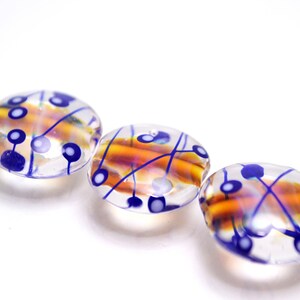 Pu&ograve; includere: Tre perline di vetro con una base trasparente e disegni blu e arancioni. Le perline hanno un motivo a spirale con linee blu e punti bianchi.