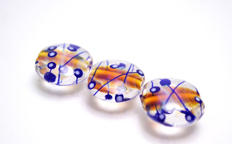 Pu&ograve; includere: Tre perline di vetro con una base trasparente e disegni blu e arancioni. Le perline hanno un motivo a spirale con linee blu e punti bianchi.