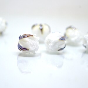 May include: White glass beads with a textured surface and iridescent blue and brown accents. The beads are arranged on a white surface.