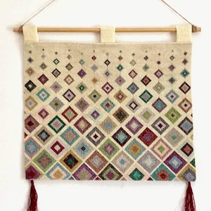 Puede incluir: Tapiz de pared tejido con un patrón geométrico de diamantes en tonos rojos, verdes, azules y morados. El tapiz es de color crema, con una barra de madera en la parte superior y borlas rojas en la parte inferior. Para decoración del hogar.