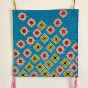 Puede incluir: Un tapiz de pared turquesa con un patrón geométrico de diamantes amarillos y rojos, y borlas rosas. El tapiz está suspendido de una varilla de madera con hilo amarillo.
