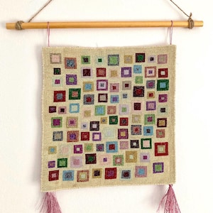 Puede incluir: Un tapiz de pared beige con un patrón repetido de cuadrados coloridos de varios tamaños. Los cuadrados son en tonos de rojo, verde, azul, morado y amarillo. El tapiz está suspendido de una varilla de madera con una cuerda y tiene borlas rosas.