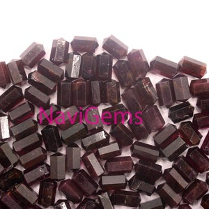 Puede incluir: Un montón de puntas de piedras preciosas de color púrpura oscuro y facetadas. Las puntas son todas de aproximadamente el mismo tamaño y forma, y están dispuestas en un patrón aleatorio. Las puntas están todas pulidas y tienen una superficie lisa y brillante.