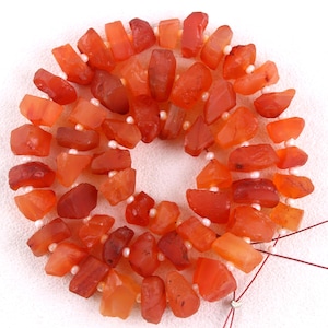 May include: A strand of carnelian beads, featuring a mix of orange and red hues, with a rough, irregular shape. The beads are strung on a white thread and are ready for jewelry making.