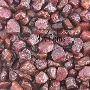 May include: A close-up view of a pile of rough, red ruby gemstones. The stones are various sizes and shapes, with a deep, rich red color. The image is taken from a slightly elevated angle, showing the texture and detail of the stones.