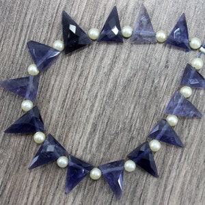 Op de afbeelding: Een ketting met driehoekige, facetgeslepen, donkerblauwe edelstenen afgewisseld met kleine, ronde, crèmekleurige parels. De edelstenen hebben een geometrische vorm en zijn geregen op een dun, lichtblauw draadje.
