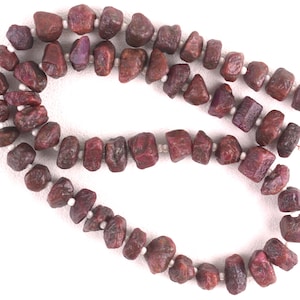 May include: A strand of deep red ruby gemstone beads, each bead is irregularly shaped and has a rough, natural finish. The beads are strung on a white thread.