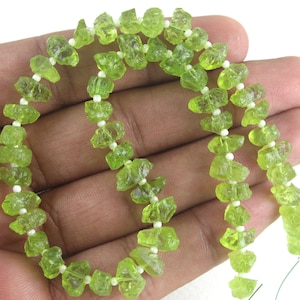 Puede incluir: Una hebra de cuentas de peridoto verde, cada cuenta tiene una forma irregular y rugosa. Las cuentas est&aacute;n enhebradas en un hilo blanco con peque&ntilde;as cuentas blancas entre cada cuenta de peridoto.