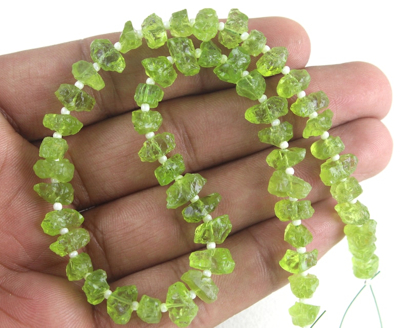 Puede incluir: Una hebra de cuentas de peridoto verde, cada cuenta tiene una forma irregular y rugosa. Las cuentas est&aacute;n enhebradas en un hilo blanco con peque&ntilde;as cuentas blancas entre cada cuenta de peridoto.
