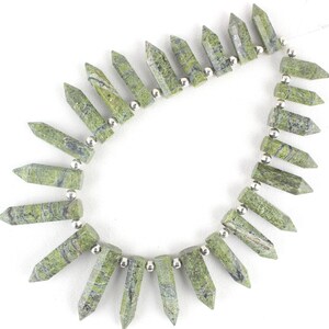 May include: A strand of green serpentine gemstone beads, each bead is a pointed, faceted, teardrop shape. The beads are strung on a silver wire with silver spacer beads between each gemstone bead.
