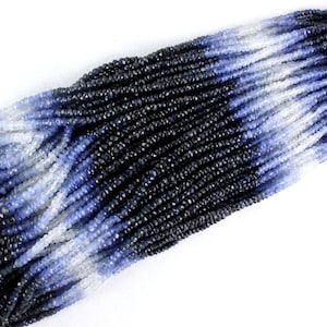 Può includere: Un filo di perle di zaffiro sfaccettate in tonalità di blu, dal scuro al chiaro, con alcune perle bianche intervallate. Le perle sono piccole e uniformi nella dimensione, e il filo è lungo e pieno.