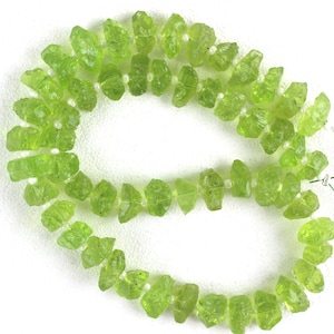 Puede incluir: Una hebra de cuentas de peridoto verde, cada cuenta es un cristal natural en bruto. Las cuentas est&aacute;n enhebradas en un hilo blanco.
