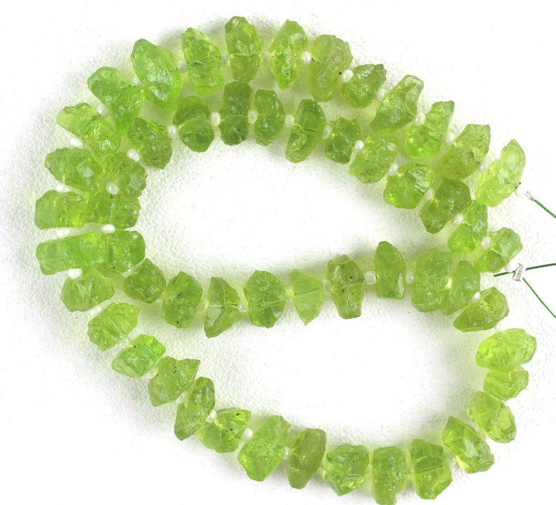 Puede incluir: Una hebra de cuentas de peridoto verde, cada cuenta es un cristal natural en bruto. Las cuentas est&aacute;n enhebradas en un hilo blanco.