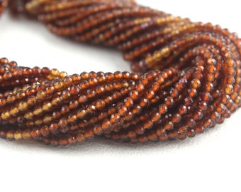May include: A close-up of strands of small, faceted, reddish-brown beads. The beads are tightly strung together, forming a twisted rope-like shape. The beads have a sparkling, polished appearance, against a white background.