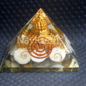 Puede incluir: Una pirámide de cristal transparente con una aguja de metal dorado y una espiral de alambre de cobre en su interior. La pirámide está llena de conchas blancas y escamas doradas.
