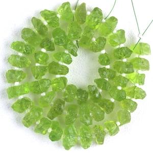 Puede incluir: Una hebra de cuentas de piedras preciosas verdes, cada cuenta tiene una forma rugosa e irregular. Las cuentas est&aacute;n enhebradas en un cord&oacute;n blanco con peque&ntilde;as cuentas blancas entre cada piedra preciosa.