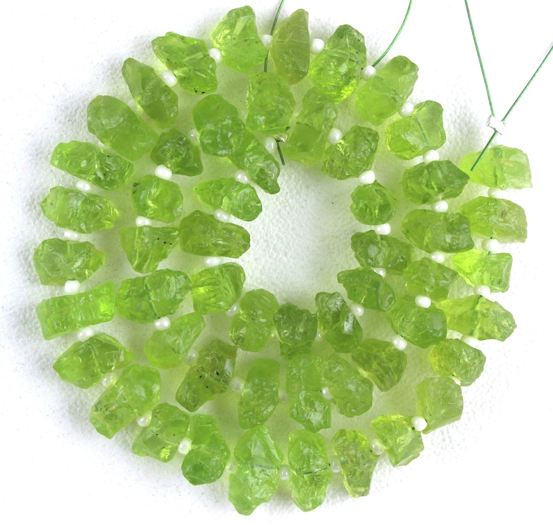 Puede incluir: Una hebra de cuentas de piedras preciosas verdes, cada cuenta tiene una forma rugosa e irregular. Las cuentas est&aacute;n enhebradas en un cord&oacute;n blanco con peque&ntilde;as cuentas blancas entre cada piedra preciosa.