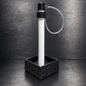 May include: A black square base with a central white vertical cylinder and a black cap. A thin, clear, flexible plastic tube connects the top of the cylinder to the side. The base features embossed decorative symbols on its front-facing side.