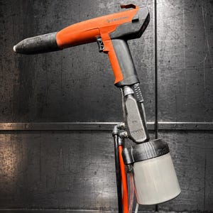May include: An orange and black powder coating spray gun labeled Electron, featuring a black handle, a trigger mechanism, and a translucent white powder container at the base with black tubing and cables extending downward from the unit.