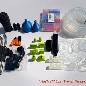 Puede incluir: Un surtido de accesorios para aspiradora, que incluye un mango negro, embudos azules y un tubo de plástico transparente. También se incluyen varias boquillas de colores, bloques verdes y un recipiente con puntas de colores. El texto "Angle Kit Only Works On Large Vac" es visible.