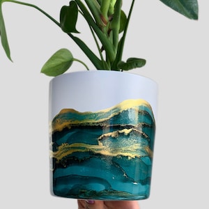Könnte beinhalten: Eine grüne Pflanze mit großen Blättern in einem weißen Keramiktopf mit einem abstrakten Design in Teal und Gold.