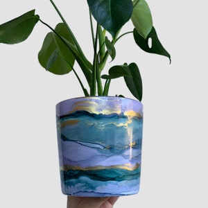 Könnte beinhalten: Eine grüne Pflanze mit großen Blättern in einem lila und blauen Topf mit goldenen Akzenten. Der Topf hat ein marmoriertes Design.