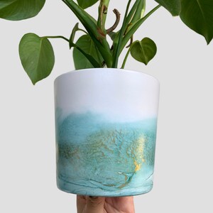May include: A white ceramic planter with a teal and gold ombre design. The planter is filled with a green leafy plant.