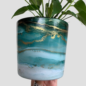 May include: A green and gold marbled ceramic planter with a houseplant growing inside. The planter has a white base with green and gold marbling.