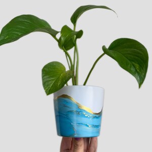 Peut inclure: Un petit pot blanc avec un motif abstrait bleu et or. Le pot est rempli d'une plante verte aux grandes feuilles.
