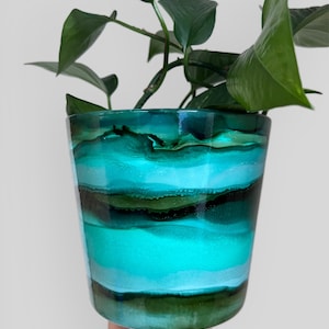 May include: A cylindrical plant pot with a layered design in teal, turquoise, and dark green. The pot contains a green leafy plant. The pot has a glossy finish. The pot's design is reminiscent of ocean waves.