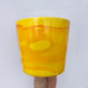 Könnte beinhalten: Eine gelbe Keramiktasse mit einem marmorierten Glasurmuster. Die Tasse hat eine abgerundete Form und eine glatte, glänzende Oberfläche.