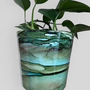 Puede incluir: Una maceta decorativa de vidrio con un diseño abstracto en tonos verdes, turquesas y marrones. La maceta contiene una planta verde con hojas. El vidrio tiene un acabado brillante y una forma cilíndrica.