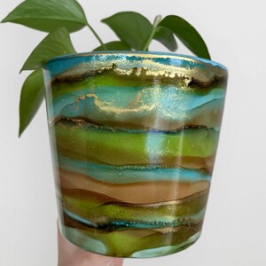 May include: A green, blue and gold striped ceramic planter with a plant inside. The planter has a glossy finish and is handmade.