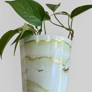 May include: A ceramic plant pot with a light green and gold marbled design. The pot contains a green leafy plant. The pot has a cylindrical shape and a gold rim. The plant pot is suitable for indoor use.