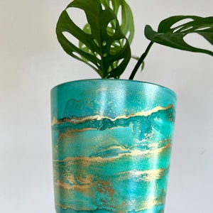 Puede incluir: Una maceta de cerámica verde azulada con detalles de pan de oro. La maceta tiene un efecto marmoleado y contiene una planta verde con hojas grandes.