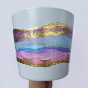 May include: A light grey ceramic planter with a gold and purple abstract design. The design features a wavy pattern with gold accents.