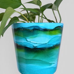 May include: A hand-painted ceramic plant pot with a glossy finish. The pot features a watercolour design in shades of turquoise, teal, and green, with gold accents. The pot is filled with green leafy plants.