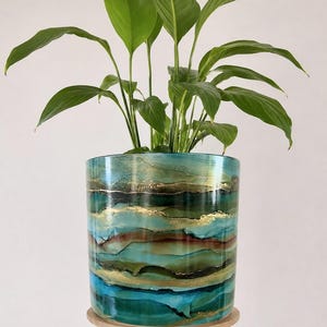 May include: A cylindrical ceramic planter featuring an abstract alcohol ink design in shades of teal, turquoise, deep green, and brown, accented with horizontal gold metallic veins. A green leafy plant grows from the top of the glossy container.