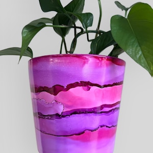 May include: A vibrant, hand-painted ceramic plant pot in shades of pink and purple. The pot features an abstract design with layered color bands. Green plant leaves and stems emerge from the top of the pot, adding a natural contrast to the colorful design.