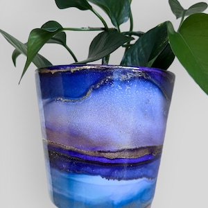 May include: A decorative ceramic plant pot with a gradient of blues and purples, accented with gold. The pot holds a green leafy plant. The pot's design features a layered effect, with the colours blending into each other. The pot is held by a hand.