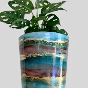 May include: A teal and gold painted ceramic planter with a green Monstera plant inside.