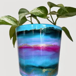 May include: A colourful, hand-painted ceramic plant pot with a layered design in shades of blue, purple, and pink, with gold flecks. The pot contains a green plant with variegated leaves. The pot is held by a hand against a white background.