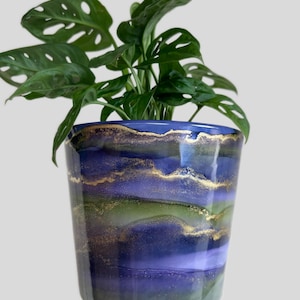 May include: A houseplant in a decorative ceramic pot. The pot is blue, green, and gold with a glossy finish and horizontal bands of colour. The plant has vibrant green leaves. The pot's rim is gold.