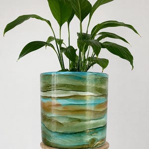 May include: A cylindrical plant pot featuring a fluid, marbled pattern in shades of turquoise, olive green, and metallic gold. A vibrant green leafy plant with long, pointed leaves grows out of the top of the container, which sits on a light wood stand.