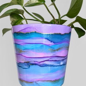 May include: A decorative plant pot with a hand-painted abstract design. The pot features bands of blue, purple, and pink, with gold metallic accents. A green leafy plant is growing from the pot. The pot is made of ceramic.