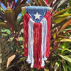 May include: A macrame wall hanging in the colors of the Puerto Rican flag. The top section is blue, with a white star in the center. Below are alternating red and white vertical strands, creating a flag-like design.