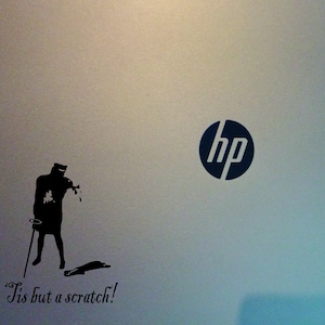Pode incluir: Um laptop prateado com um logotipo HP preto e um adesivo preto e branco de um cavaleiro que diz "Tis but a scratch!"