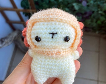 Peut inclure: Un petit jouet en peluche crocheté avec un corps blanc, une tête et des bras de couleur pêche et un chapeau rouge et pêche. Le jouet a des yeux noirs et un petit nez noir.