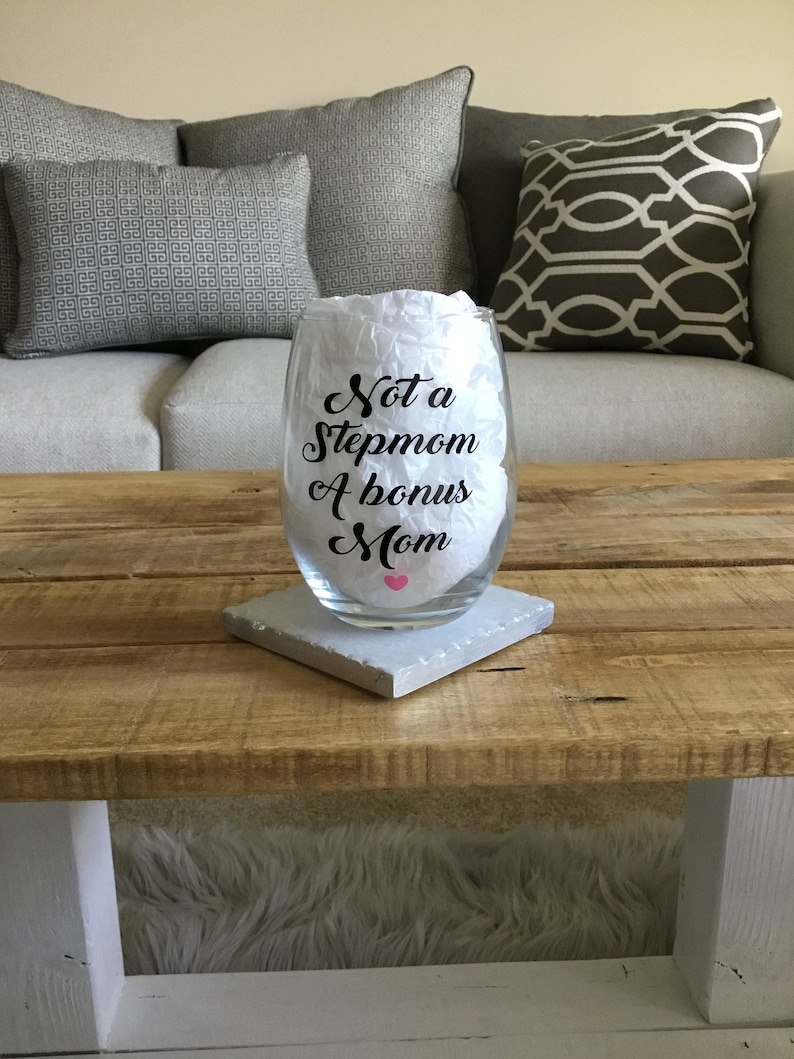 May include: A clear glass wine glass with a black vinyl decal that reads "Not a Stepmom A bonus Mom" with a pink heart. The glass is sitting on a white coaster on a wooden table.